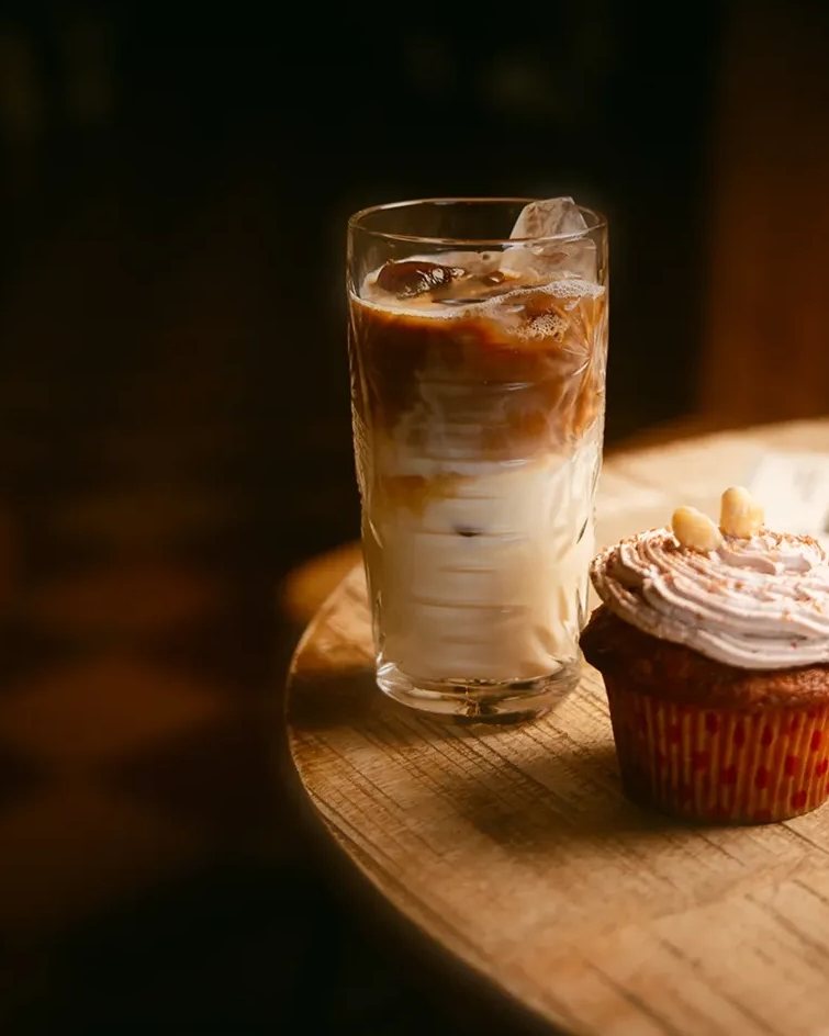 Café gelado com leite num copo alto e um cupcake com cobertura, numa mesa redonda de madeira