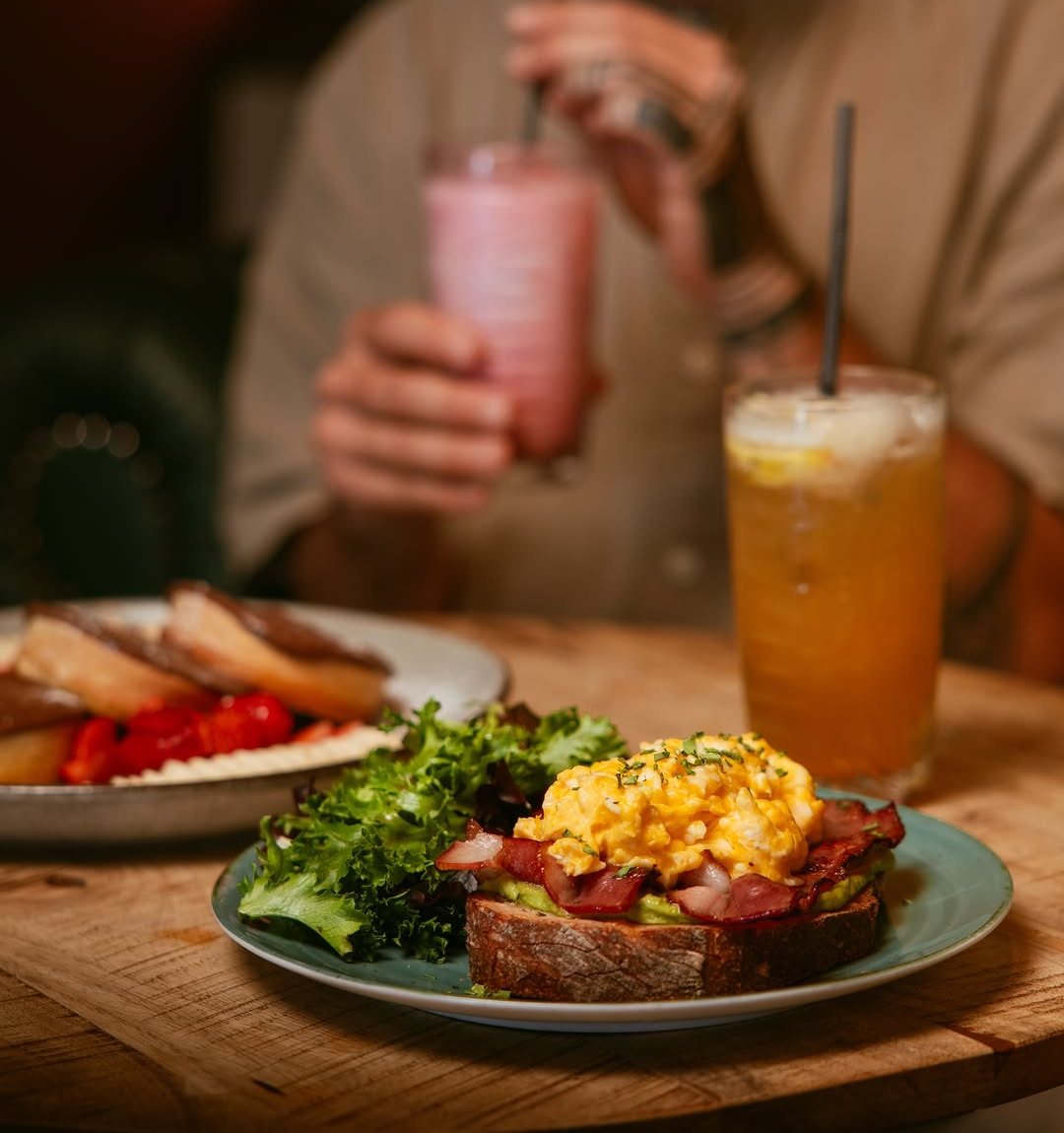 Tosta com abacate, bacon e ovos mexidos num prato azul, ao lado de salada, com um sumo cor de laranja e um batido cor-de-rosa ao fundo