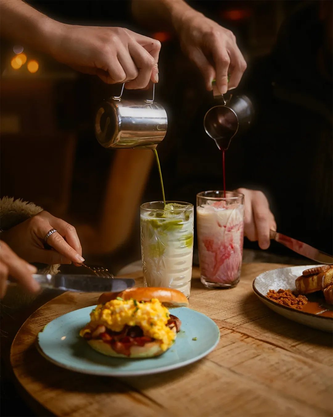 Bagel com ovos mexidos e bacon num prato azul, enquanto duas bebidas geladas são servidas na mesa de madeira